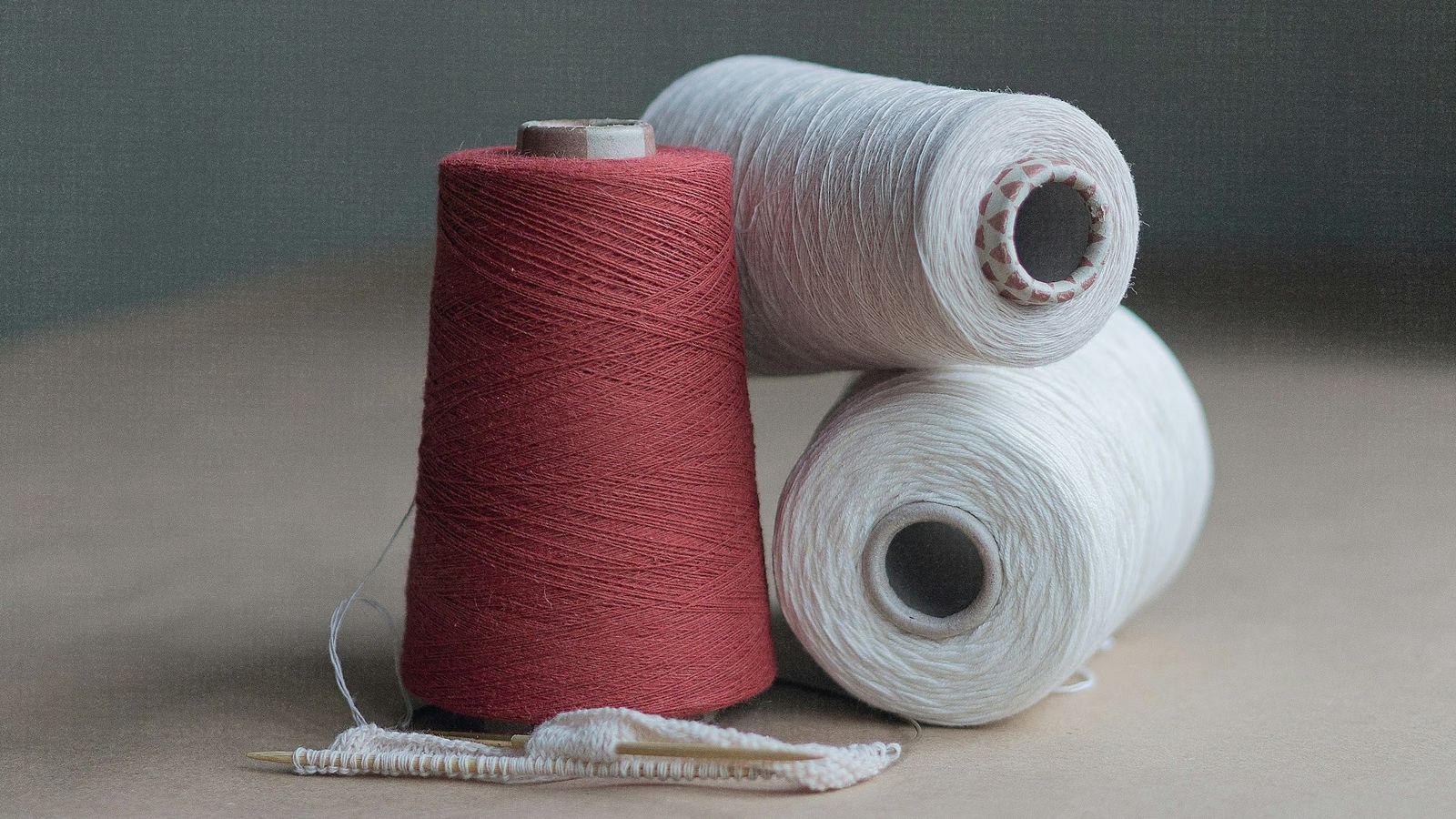 Red and white cotton yarn cones beside a hand-knitted swatch on needles